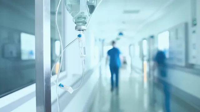 Prim-plan cu perfuzie și tub transparent; fundal neclar cu coridor și două siluete albastre.