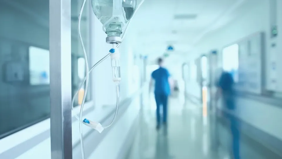 Prim-plan cu perfuzie și tub transparent; fundal neclar cu coridor și două siluete albastre.