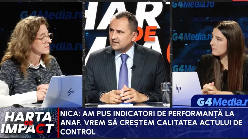 Un bărbat cu cravată purtată într-un costum închis la culoare stă la o masă între două femei în cadrul unui studio de televiziune.