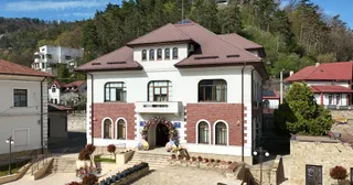 O clădire albă cu acoperiș maro și detalii din cărămidă roșie este situată în fața unui deal împădurit sub un cer senin.