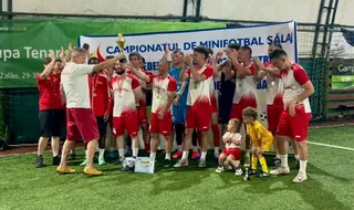 Un grup de bărbați în echipamente sportive roșii și albe stau pe un teren de fotbal artificial lângă un banner alb și un trofeu auriu.
