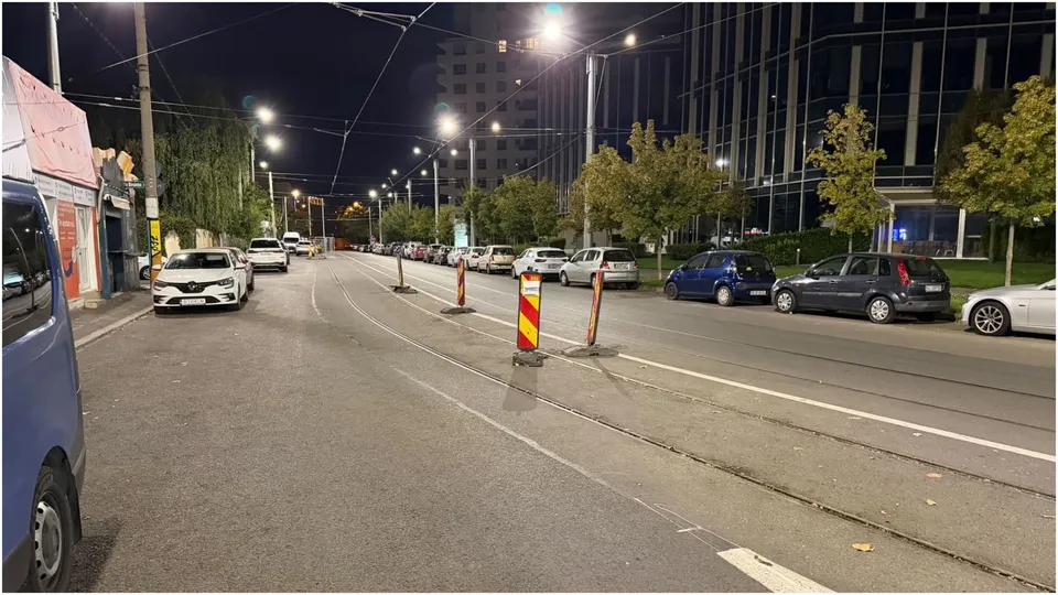 Patru balize rutiere sunt așezate pe asfaltul unei străzi urbane, lângă mașini parcate și clădiri sub iluminarea nocturnă.