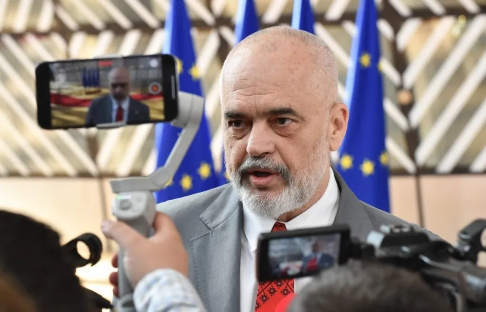 Un bărbat cu barbă căruntie și sacou gri vorbește în timp ce un telefon mobil și o cameră video îi încadrează fața.