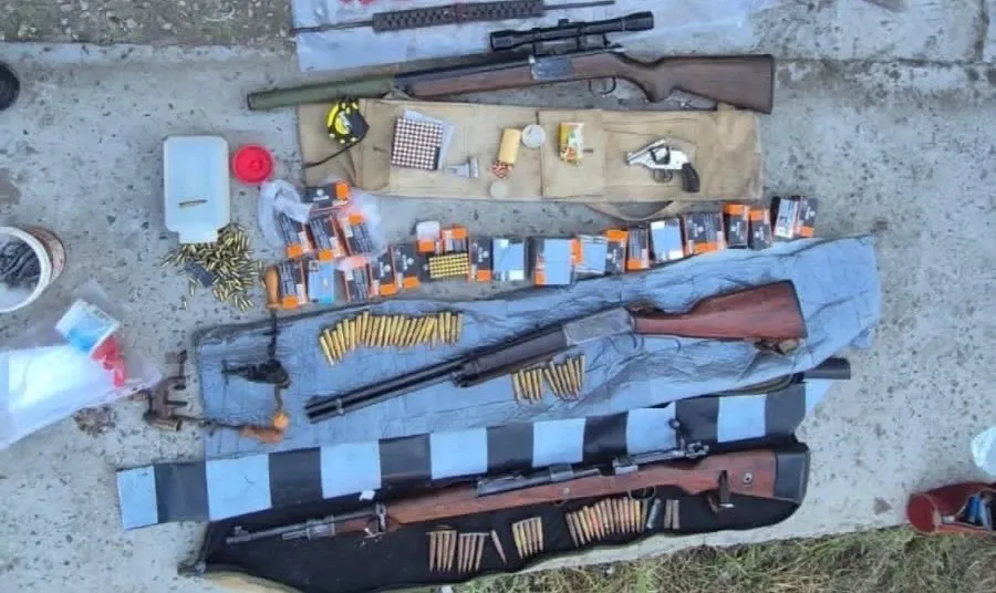 O imagine de sus a mai multor arme de foc, inclusiv puști și un pistol, alături de o cantitate mare de muniție, așezate pe o suprafață de beton.