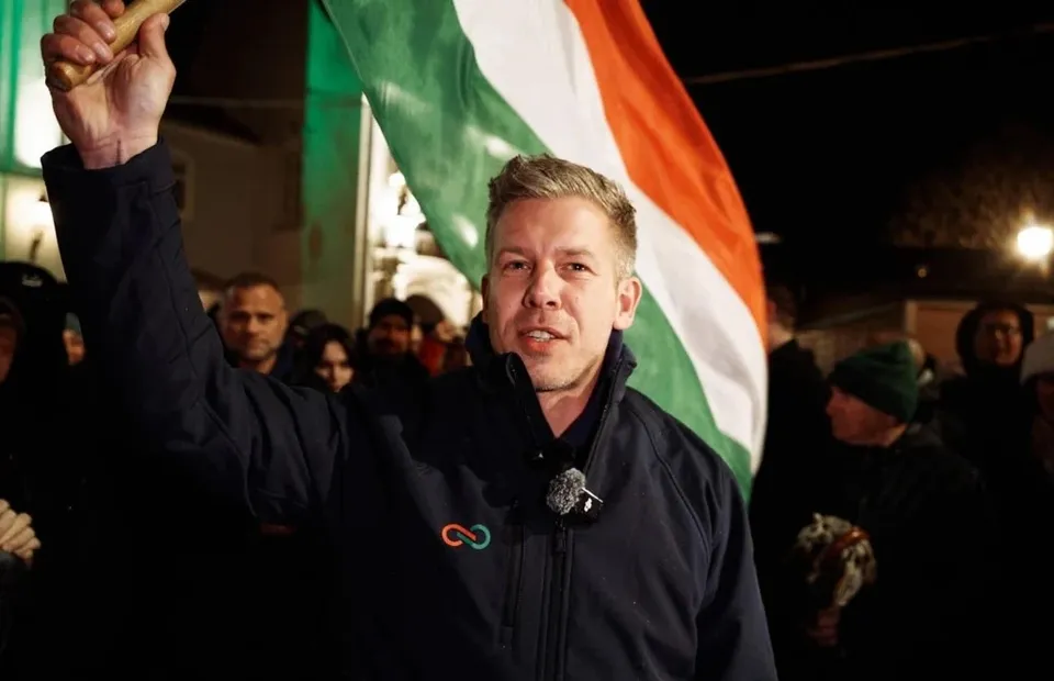 Un bărbat cu părul scurt și o jachetă neagră ridică un mâner de lemn în fața unui steag tricolor sub un cer de noapte.