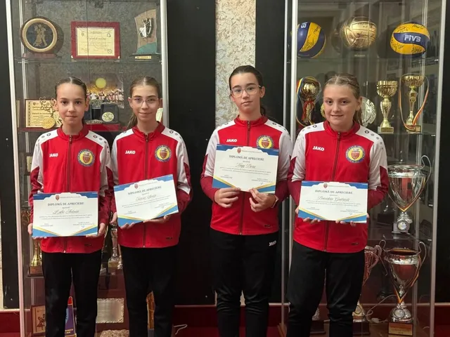 Patru tinere poartă jachete roșii și negre, ținând diplome albe în fața unui vitrină plină cu trofee și mingi de sport.