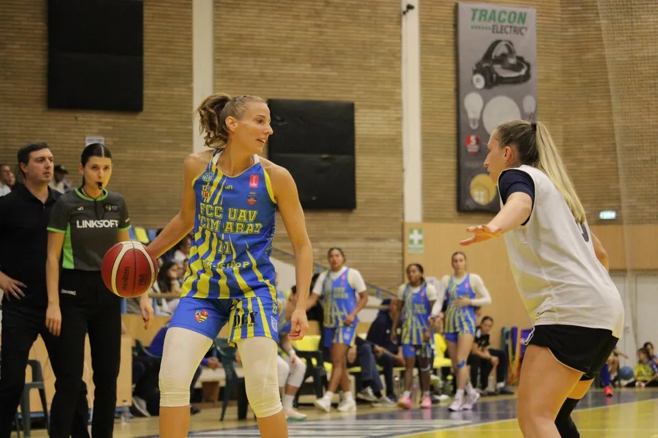 O jucătoare de baschet în echipament albastru și galben driblează mingea pe un teren de sport, în fața unui arbitru și a altor jucătoare.