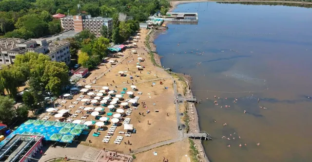 O vedere aeriană a unei zone de plajă cu numeroase umbrele de parasol aliniate și persoane care înoată într-un lac mare.