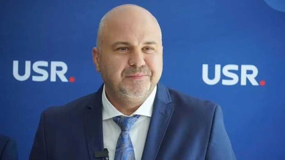 Un bărbat cu părul scurt și barbă stufoasă poartă sacou albastru, cămașă albă și cravată cu modele, stând în fața unui fundal albastru cu logouri albe.