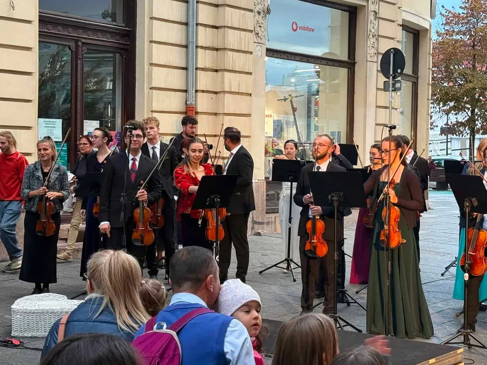Un grup de muzicieni cu instrumente de coarde cântă pe o stradă lângă clădiri, în fața unui public.