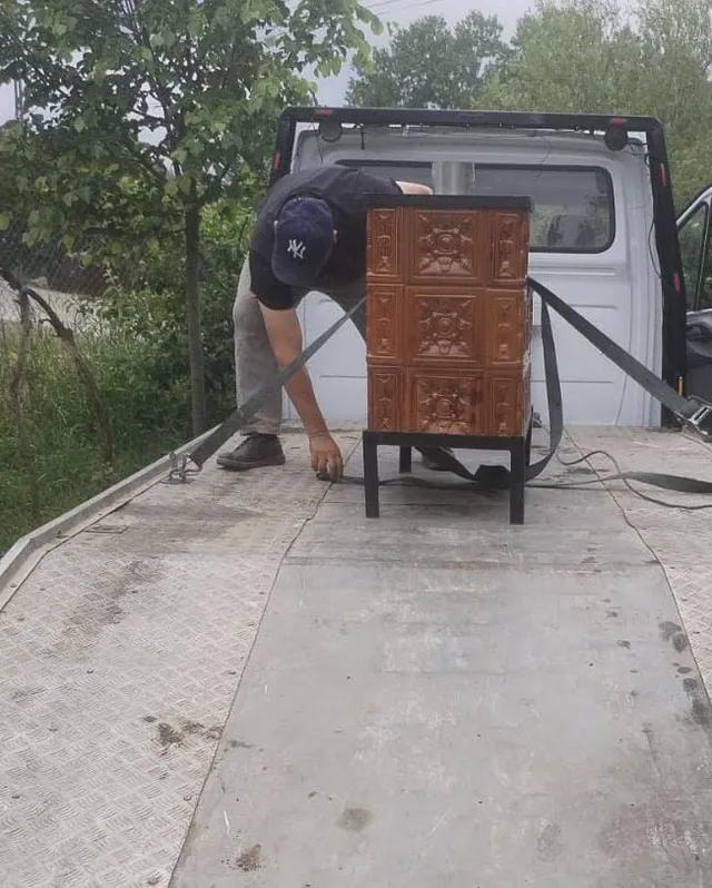 Un bărbat cu șapcă și pantaloni deschiși stă în spatele unui autovehicul alb, lângă un obiect maro dreptunghiular fixat cu curele de prindere.