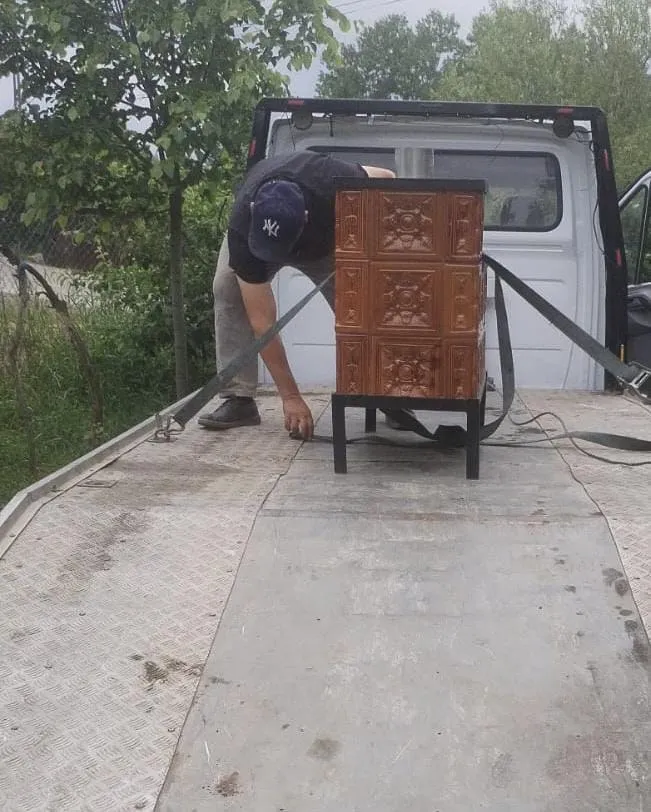 Un bărbat cu șapcă și pantaloni deschiși stă în spatele unui autovehicul alb, lângă un obiect maro dreptunghiular fixat cu curele de prindere.
