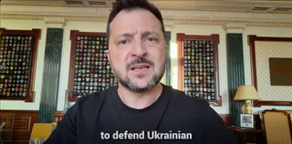 Un bărbat cu barbă și păr scurt, îmbrăcat într-un tricou negru, privește spre cameră într-o încăpere cu panouri decorative pe perete și o lampă pe o masă.