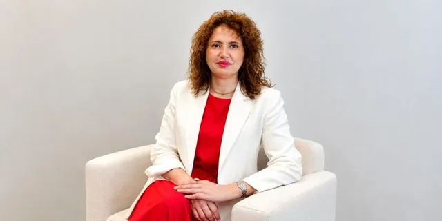 O femeie cu părul creț, purtând un sacou alb peste o rochie roșie, stă așezată într-un fotoliu de culoare deschisă.