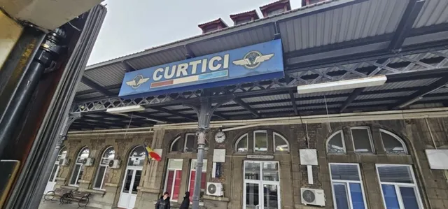 Un panou albastru cu textul CURTICI este suspendat sub o structură metalică de acoperire deasupra unei clădiri cu ferestre și unități de aer condiționat.