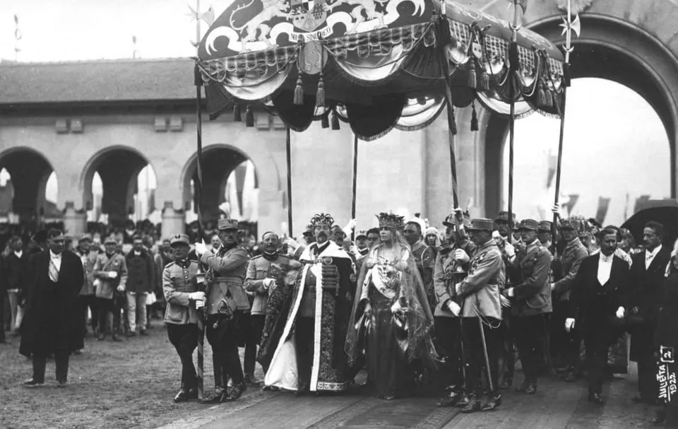 Un grup de oameni în uniforme și haine formale stau sub un baldachin decorat, în fața unor arcade de piatră.