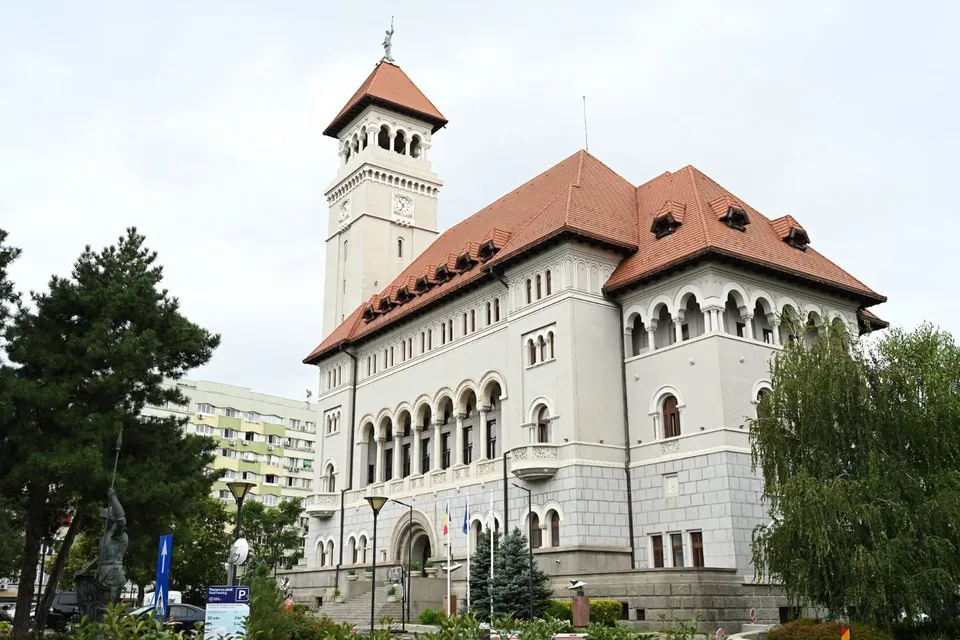O clădire albă cu acoperiș roșu și un turn înalt este înconjurată de copaci verzi sub un cer înnorat.