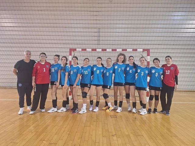 O echipă de handbal formată din femei și un bărbat stă în linie în fața unei porți de handbal pe un teren de lemn.