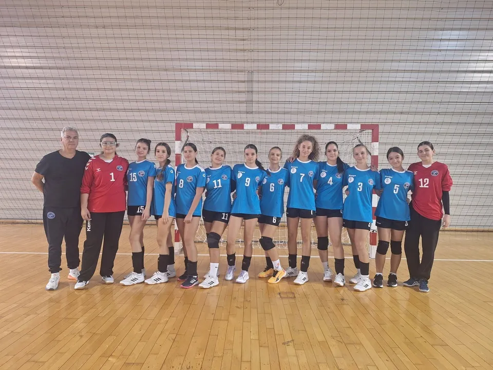 O echipă de handbal formată din femei și un bărbat stă în linie în fața unei porți de handbal pe un teren de lemn.