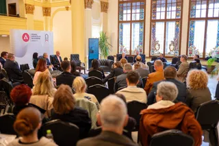 Oamenii stau așezați pe scaune negre într-o sală cu ferestre înalte și decorate, privind spre un podium cu un panou alb.