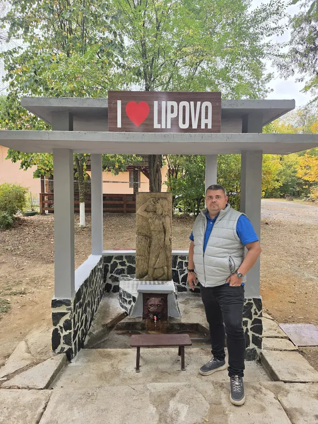 Un bărbat stă lângă un izvor de apă acoperit de o structură de beton cu un panou pe care scrie Lipova.