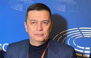 Un bărbat cu păr scurt și scurt, îmbrăcat într-un sacou albastru și un tricou negru, stă în fața unui fundal albastru cu embleme albe.