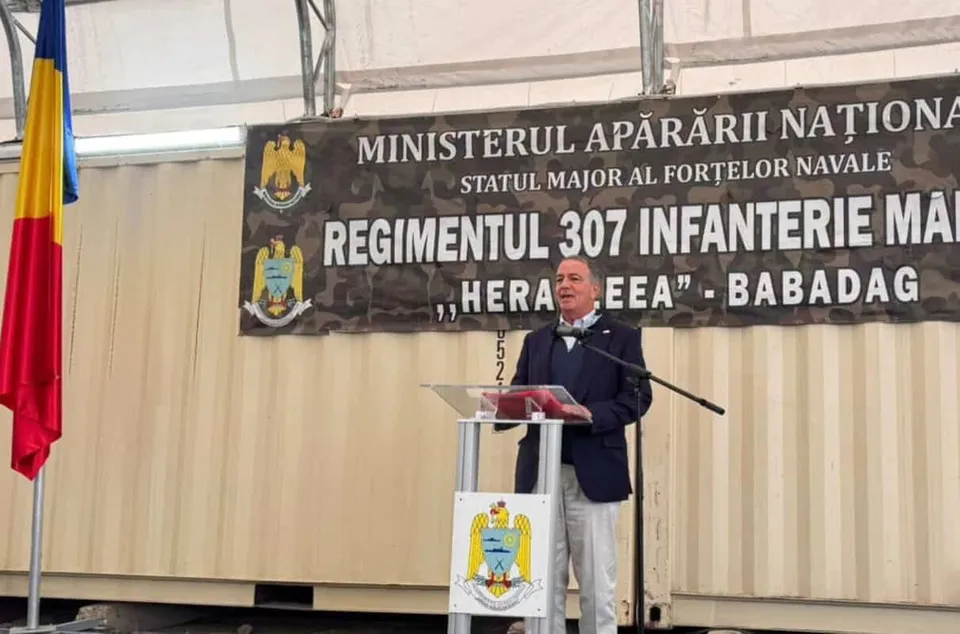 Un bărbat într-un costum închis vorbește la un pupitru aflat în fața unui panou mare cu inscripții și embleme.