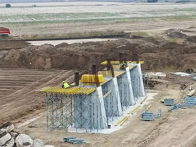 Structuri de beton gri cu piloni de susținere sunt încadrate în schele metalice pe un teren de construcție cu pământ și utilaje.