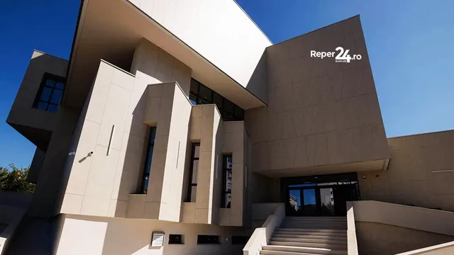 O clădire modernă, cu fațade din beton și ferestre înalte, sub un cer senin, cu o scară largă ce duce la intrare.
