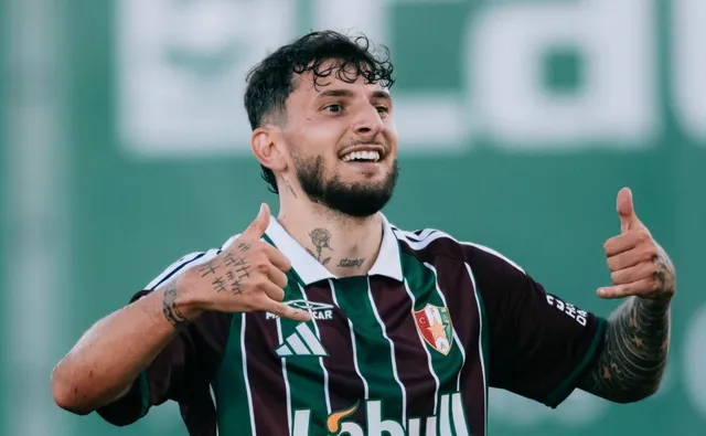 Un bărbat cu barbă și tatuaje pe brațe poartă un tricou sportiv în culorile maro și verde, făcând gestul cu degetul mare în sus.