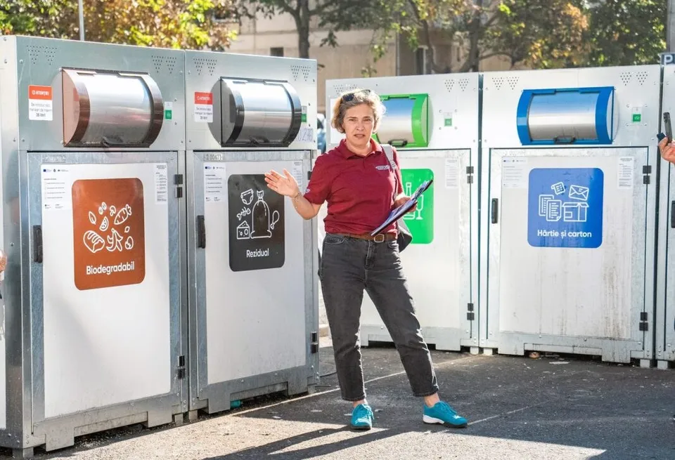 O femeie cu tricou roșu și pantaloni gri stă lângă containere de reciclare de diferite culori pe un trotuar.