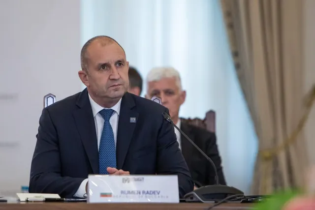 Un bărbat cu părul scurt și costum închis la culoare stă la o masă cu un microfon și un semn de identificare în față.