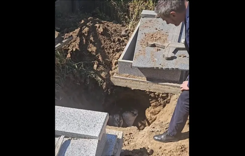 Un bărbat se apleacă și privește într-o groapă săpată în pământ, lângă o construcție funerară din piatră. În groapă se observă un sac alb.