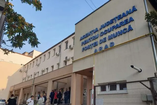 O grupă de persoane stă la intrarea într-o clădire albă pe care este scris cu litere albastre Agenția Județeană pentru Ocuparea Forței de Muncă Arad.