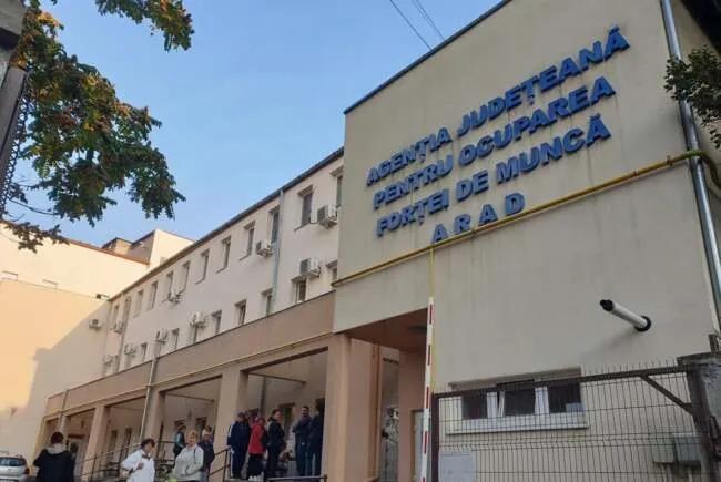 O grupă de persoane stă la intrarea într-o clădire albă pe care este scris cu litere albastre Agenția Județeană pentru Ocuparea Forței de Muncă Arad.