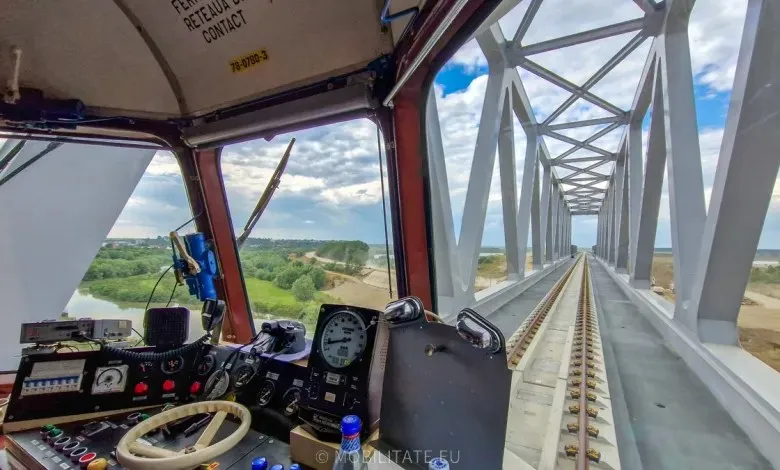 Privire de pe interiorul unei locomotive spre șinele de pe un pod metalic cu structură albă sub un cer cu nori.