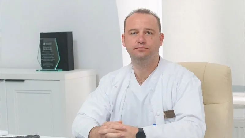 Un bărbat cu halat alb stă așezat la un birou într-o încăpere cu un dulap alb și un trofeu în fundal.