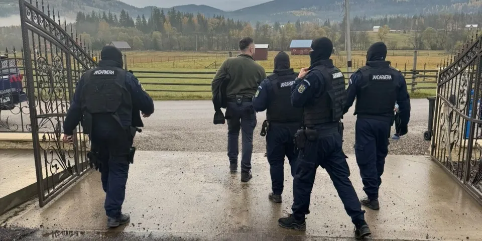 Cinci persoane purtând uniforme închise și echipamente tactice merg pe un drum pavat printre porți metalice și câmpuri deschise.