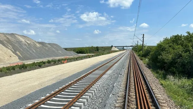 Două șine de tren cu traverse de beton și lemn se întind spre orizont sub un cer cu nori dispersate.