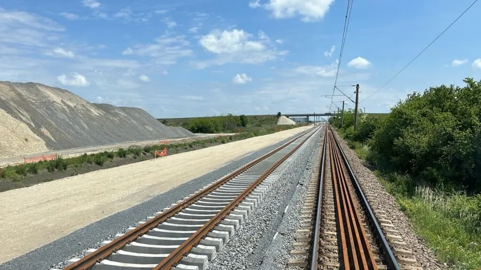 Două șine de tren cu traverse de beton și lemn se întind spre orizont sub un cer cu nori dispersate.