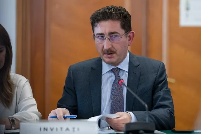 Un bărbat cu ochelari, purtând sacou închis și cravată, stă la o masă cu un microfon și ține un document și un pix.