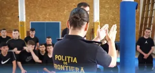 O femeie cu părul prins într-un coc ridică mâinile în poziție de aplau, purtând un tricou negru cu inscripția POLIȚIA DE FRONTIERĂ, în fața unui grup de bărbați.