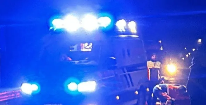 O ambulanță cu luminile de semnalizare aprinse este parcată pe un drum noaptea lângă un grup de persoane.