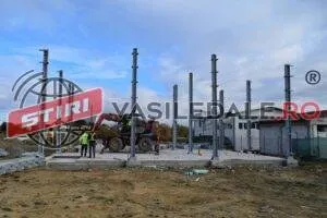 Mai multe stâlpi metalici verticali sunt montați pe o platformă de beton lângă un utilaj de construcții și doi oameni.