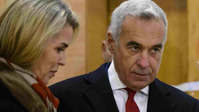Un bărbat cu părul cărunt și cravată roșie stă lângă o femeie cu părul blond și eșarfă maro.