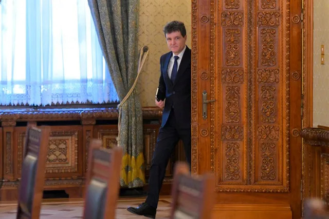 Un bărbat într-un costum închis și cravată albastră stă în picioare lângă o ușă din lemn sculptat într-o încăpere cu mobilier decorat.