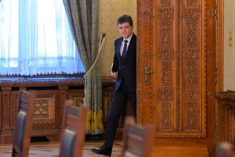 Un bărbat într-un costum închis și cravată albastră stă în picioare lângă o ușă din lemn sculptat într-o încăpere cu mobilier decorat.