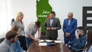 Un grup de persoane stă în jurul unei mese de lemn pe care se află foi de hârtie, în timp ce un bărbat scrie pe o foaie.