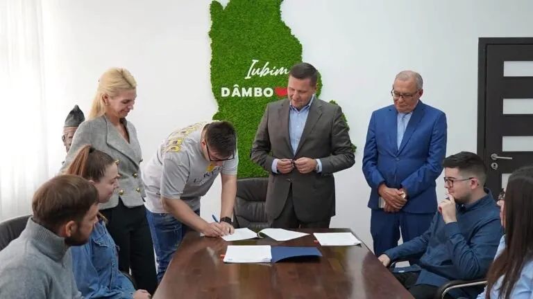 Un grup de persoane stă în jurul unei mese de lemn pe care se află foi de hârtie, în timp ce un bărbat scrie pe o foaie.