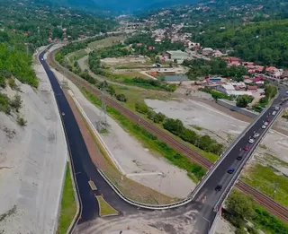 O vedere aeriană prezintă o autostradă nouă cu două benzi, o cale ferată și un peisaj cu dealuri verzi și case dispersate.